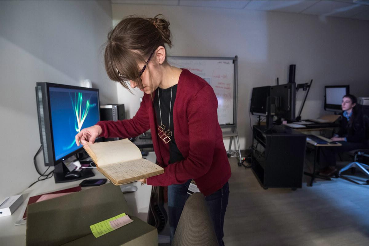 librarian in a digital lab surrounded by computer screens holds an old book.